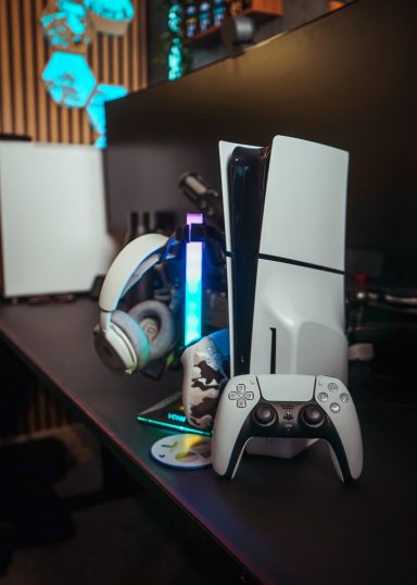 A PlayStation 5 console with accessories on a desk, illuminated by colourful lighting.