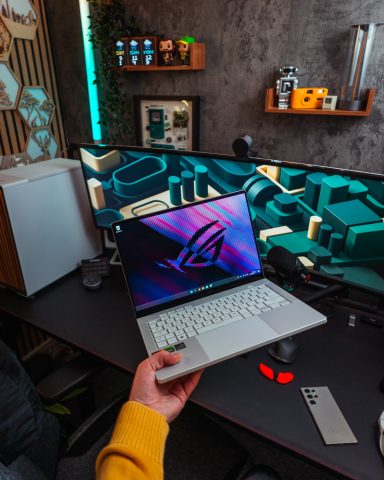 A person holds a laptop in a stylish workspace with dual monitors.